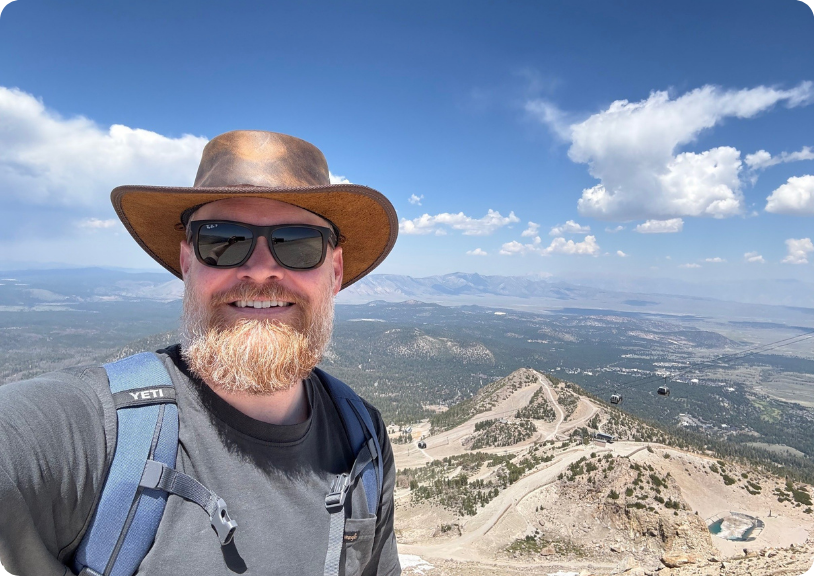 Adam stands atop a mountain, showcasing a scenic landscape under a blue sky.