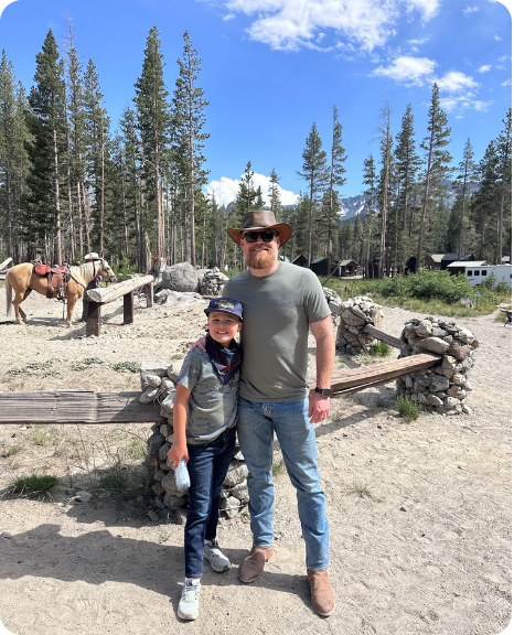 A man and a child pose together outdoors in a forested area, with horses and a rustic setting in the background.