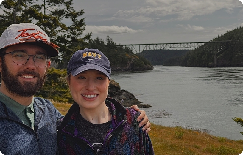 Two people smiling together by a river with a bridge in the background, surrounded by trees and a cloudy sky.