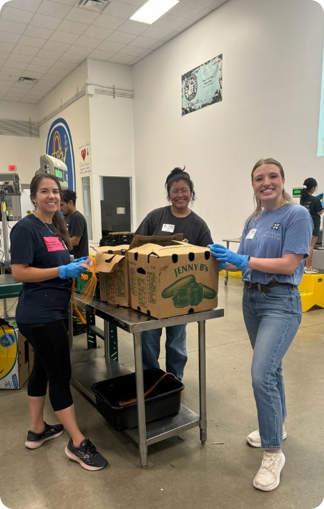 Three volunteers smile while sorting produce into boxes at a food distribution center.