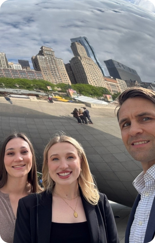 Three smiling individuals pose in front of a reflective sculpture, capturing a vibrant urban backdrop.