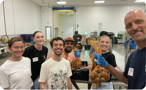 A diverse group of six volunteers smiles while holding bags of produce in a warehouse setting.