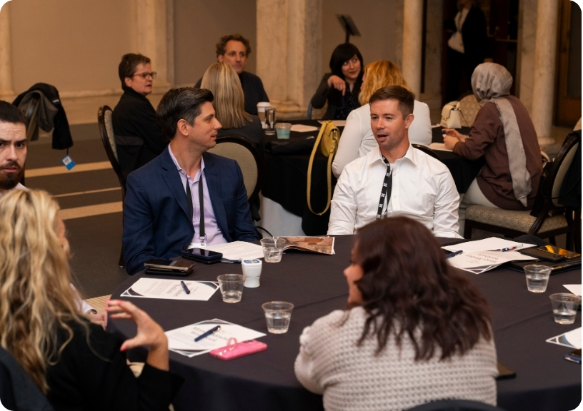 Two men engage in conversation at a round table during a networking event, with attendees in the background.