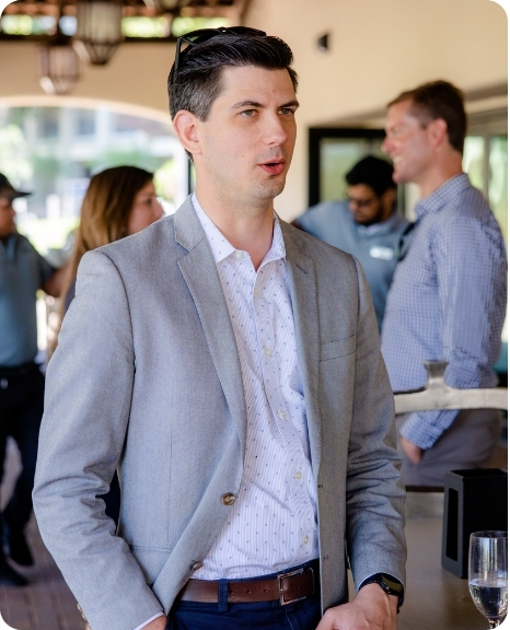 Andrew Kulha engages in conversation at a networking event, dressed in a light gray blazer and standing near a bar.