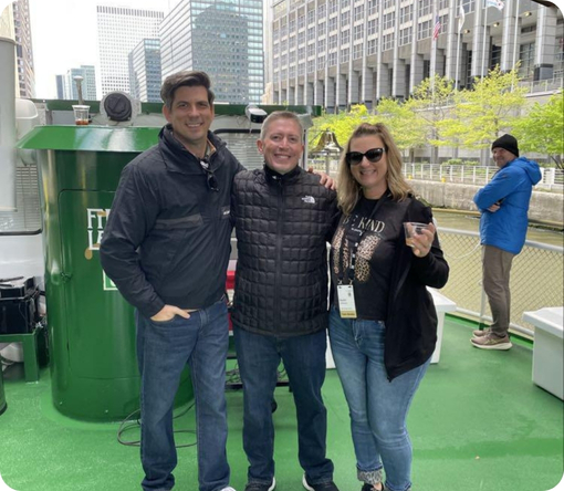 Three people pose together on a boat, with city buildings and a river in the background, enjoying a casual outing.