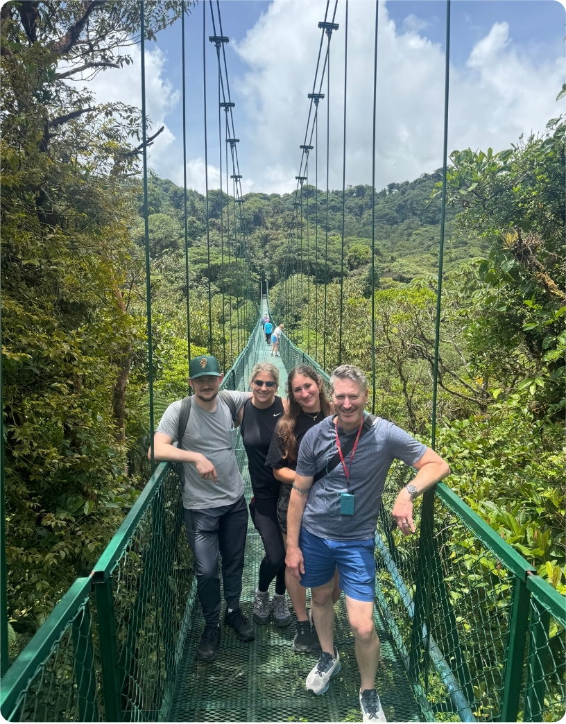 Four friends pose on a suspension bridge surrounded by lush greenery and a bright blue sky.