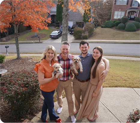 A family stands together on a porch, holding two small dogs, with autumn foliage and a suburban neighborhood in the background.