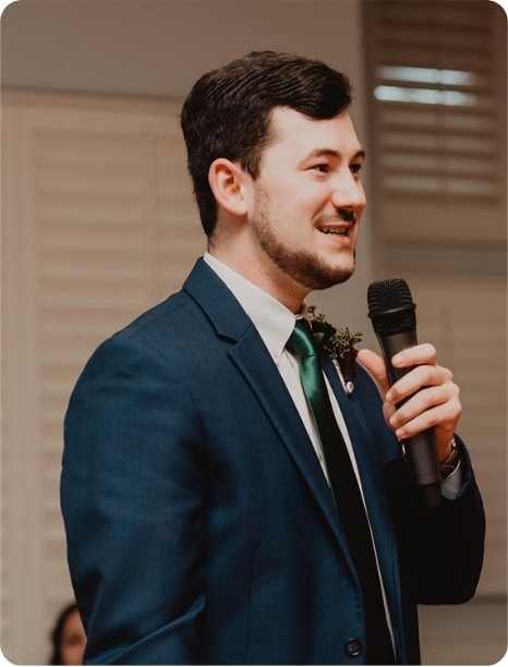 Braydan Meyers delivers a speech while holding a microphone, dressed in a formal suit with a green tie and boutonniere.