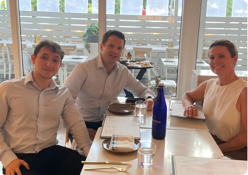 Three people sit at a dining table with menus, water glasses, and a blue bottle, enjoying a meal together.