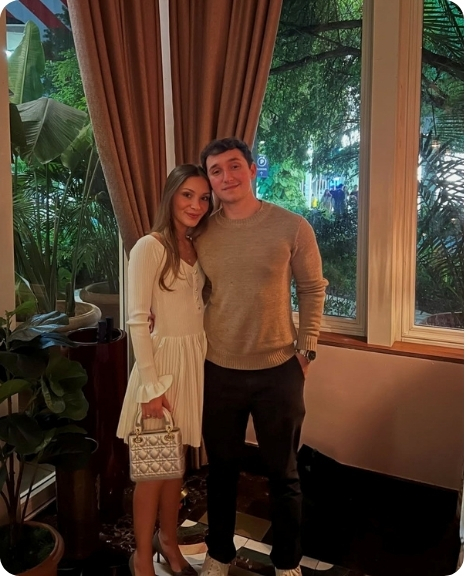A couple poses together in a cozy indoor setting, with plants and a window in the background, showcasing a warm atmosphere.