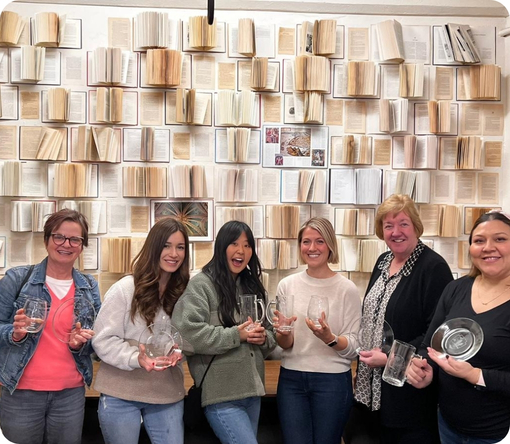 A group of six women proudly displaying glassware in front of a unique book-themed wall, celebrating Diane Williamson Mission Wealth.