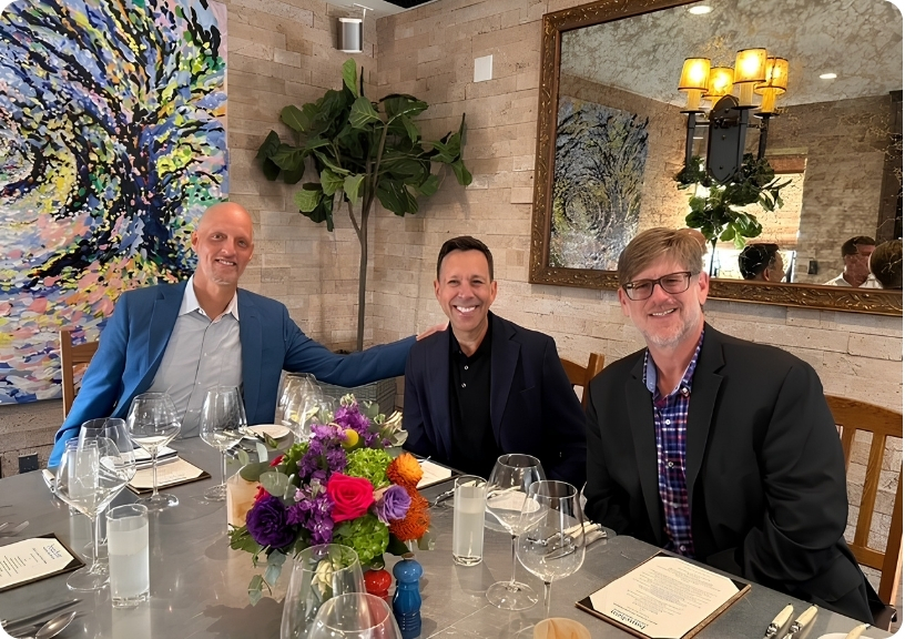 Three men sit at a dining table with a floral centerpiece, celebrating together, reflecting the camaraderie of Jeff Avila Mission Wealth.