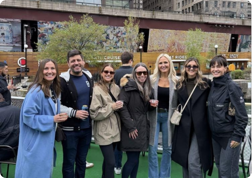 A group of seven friends smiles together on a boat, enjoying drinks with a scenic backdrop of city buildings.