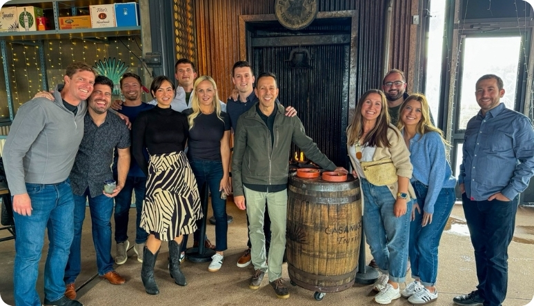 A group of friends poses together at a bar, smiling and enjoying their time, with a rustic barrel in the background.