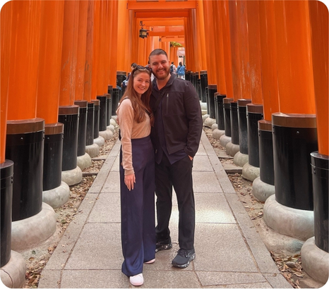 A couple stands together smiling in a vibrant pathway lined with tall orange torii gates.