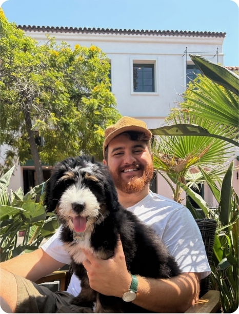 Smiling man in a hat holds a playful puppy, surrounded by lush greenery and a sunny outdoor setting.