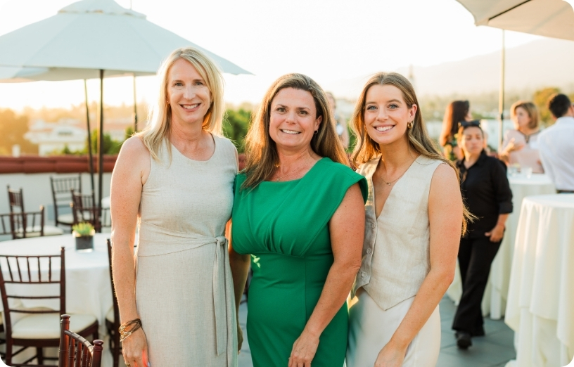 Three women pose together at a rooftop event during sunset, showcasing a lively social atmosphere.