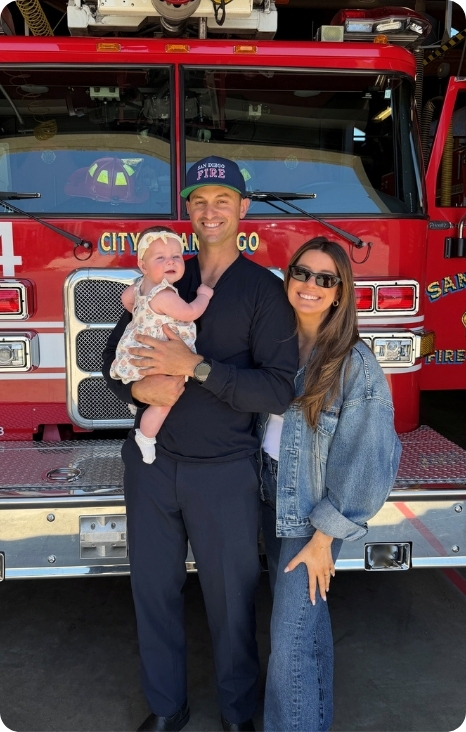 A smiling couple holds a baby in front of a fire truck, showcasing family and community connection.