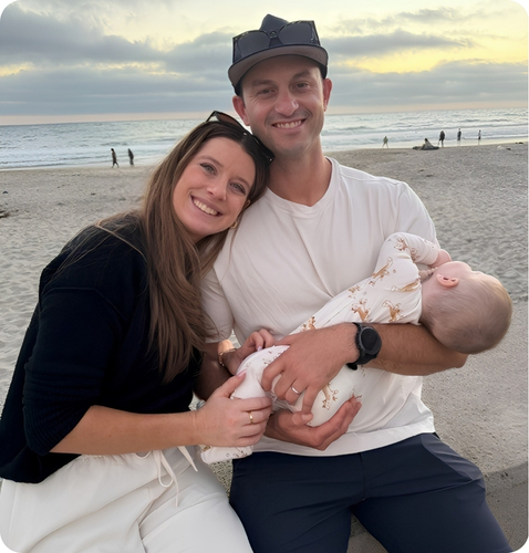 Couple smiling while holding a sleeping baby on a beach at sunset, capturing a moment of family togetherness.