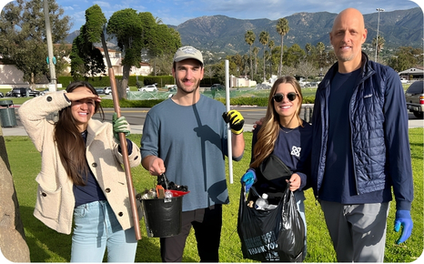 Four volunteers holding trash collection tools and bags, posing in a park with mountains in the background.