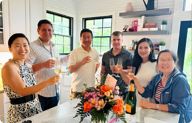 A group of six people celebrate with champagne in a bright kitchen, surrounded by flowers and cheerful expressions.