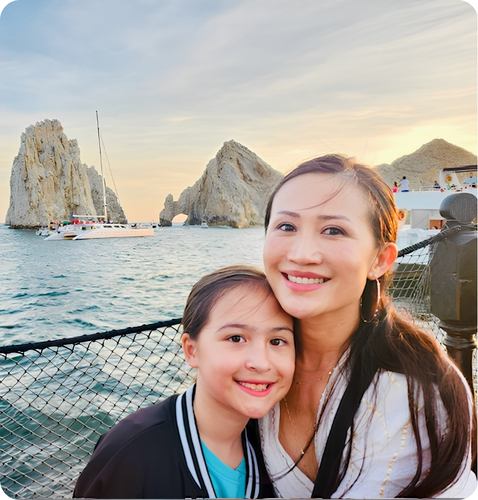 Leah Naish smiles with her child by the ocean, featuring scenic rock formations and a boat in the background.