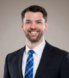 Cole Haddock smiles confidently in a professional suit and tie against a neutral background.