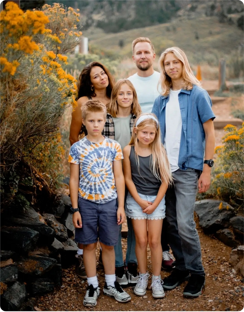 A family of six poses together outdoors, surrounded by colorful flowers and rocky terrain.
