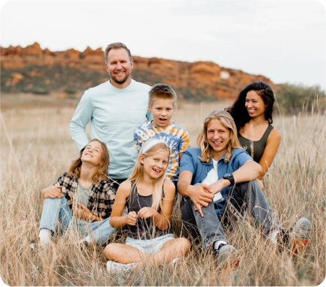 A family of six smiles together in a grassy field with rocky hills in the background.