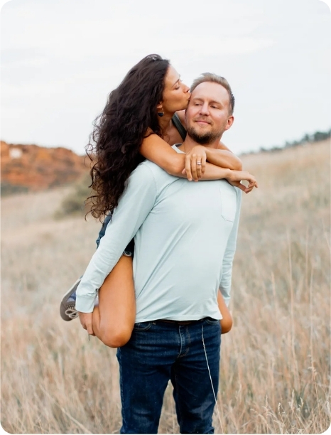 A couple shares a joyful moment outdoors, with the woman playfully kissing the man on the cheek while he carries her.