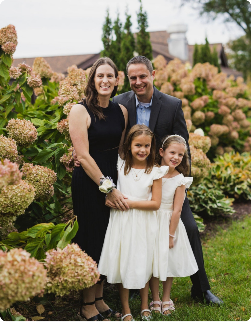 A family of four poses together in a garden, smiling in front of blooming hydrangeas, dressed for a special occasion.