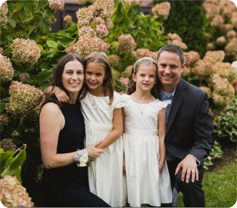 Family portrait in a garden, featuring two girls in white dresses with their parents, surrounded by blooming hydrangeas.