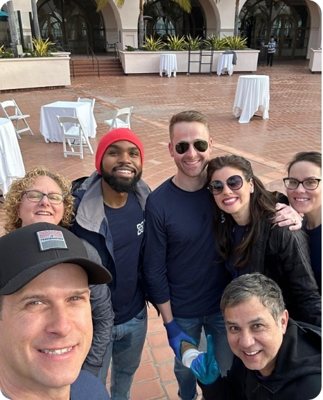 A diverse group of six smiling volunteers poses together outdoors, ready for a community service event.