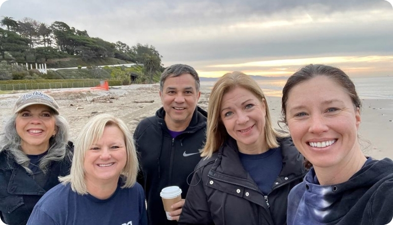 Group of five friends smiling together on a beach during sunrise, enjoying coffee and a relaxed moment outdoors.