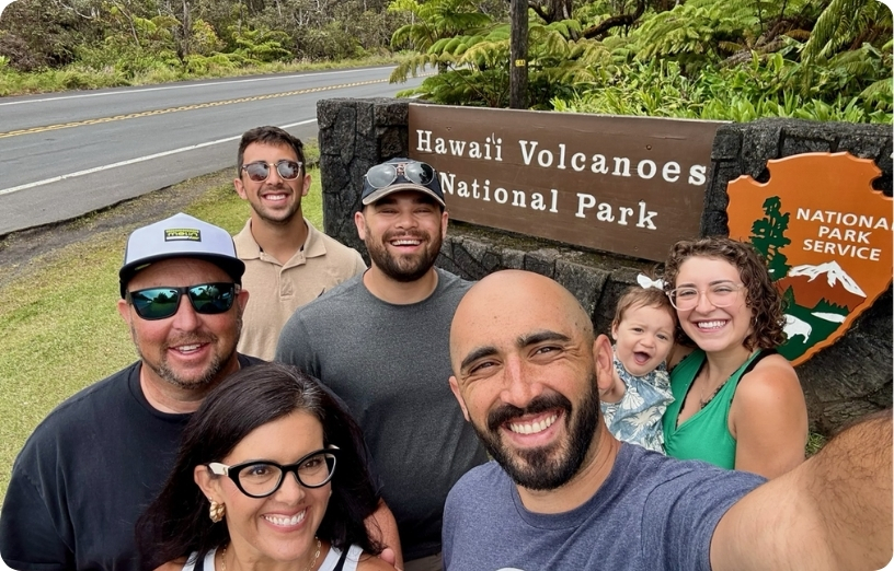 Group of six people smiling in front of the Hawaii Volcanoes National Park sign, enjoying their visit together.