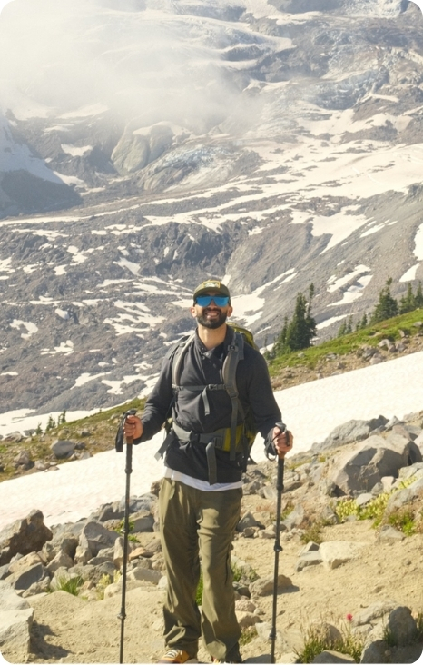 Auddie Sweet stands on a mountain trail, equipped for hiking, with a stunning landscape of snow-capped peaks behind him.