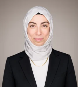 Professional portrait of Esra Dulger wearing a black blazer and light-colored hijab, smiling against a neutral background.