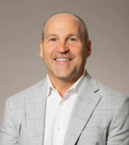 Smiling man in a light gray blazer and white shirt against a neutral background, representing Geoff Gaggs.