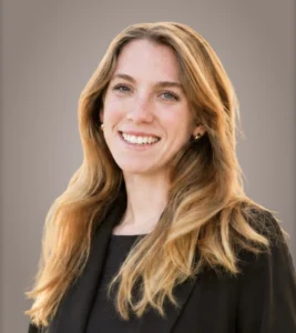 Smiling young woman with long, wavy hair wearing a black blazer against a neutral background.