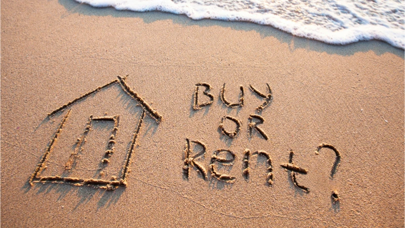 Sand writing of a house with the words "Buy or Rent?" on a beach, symbolizing housing choices.