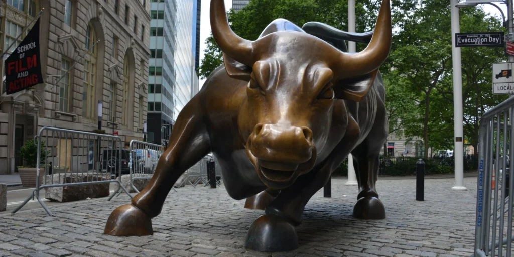 Bronze bull statue symbolizing stock market growth, located in a bustling urban area with greenery and buildings.