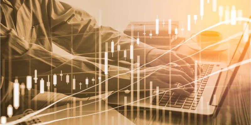 Hands typing on a laptop with overlaid stock market graphs, symbolizing financial analysis and investment trends.