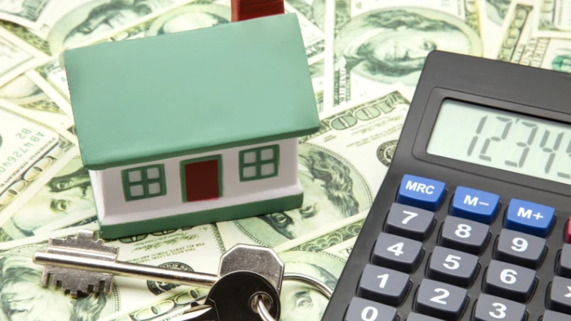 A toy house, keys, and a calculator are placed on a background of money, symbolizing financial aspects of second homes.