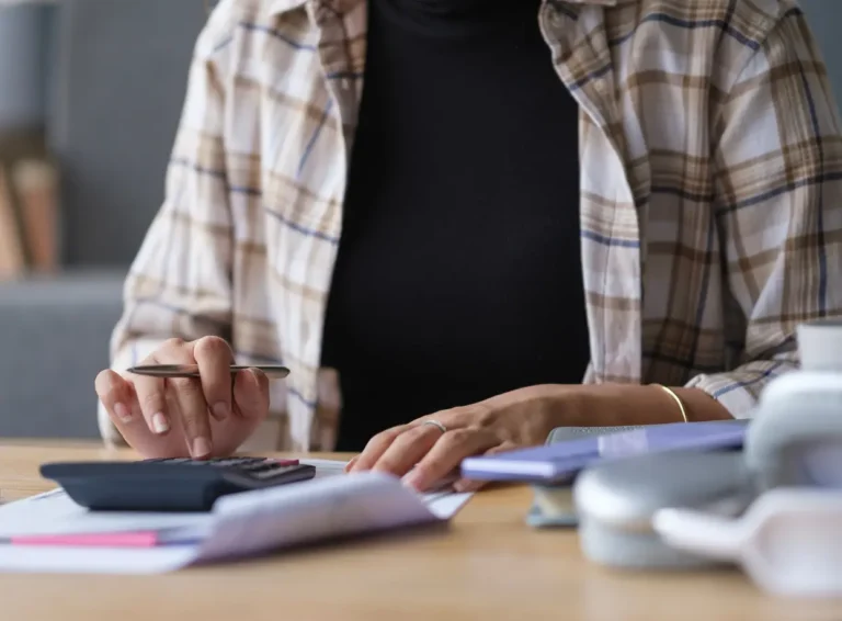 A person calculates finances with a calculator, papers, and a pen, wearing a plaid shirt and black top.