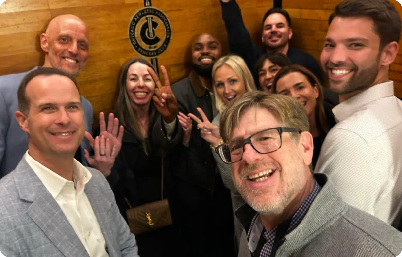 A cheerful group of professionals poses for a selfie, celebrating with smiles and peace signs in a warm, wooden setting.