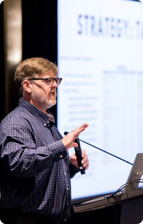 Presenter discussing strategic insights at a conference, with a slide visible in the background.