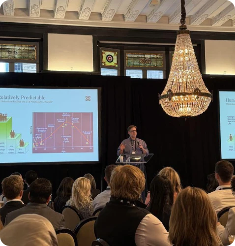 A speaker presents data on a screen during a conference, with an engaged audience in a well-lit, elegant venue.
