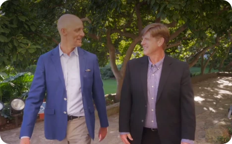 Two men walk and converse in a lush garden, showcasing a friendly and engaging atmosphere.