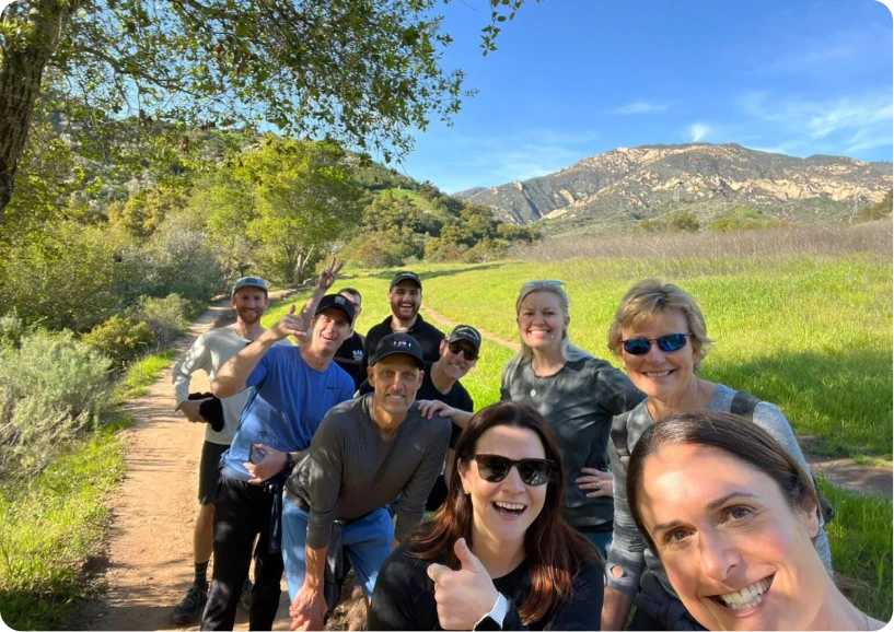 A group of happy hikers poses for a selfie on a scenic trail with mountains and greenery in the background.