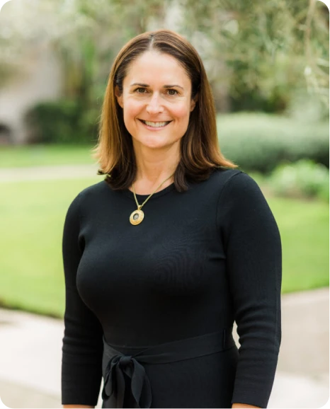 Smiling woman in a black dress stands outdoors, showcasing a friendly demeanor against a lush green background.